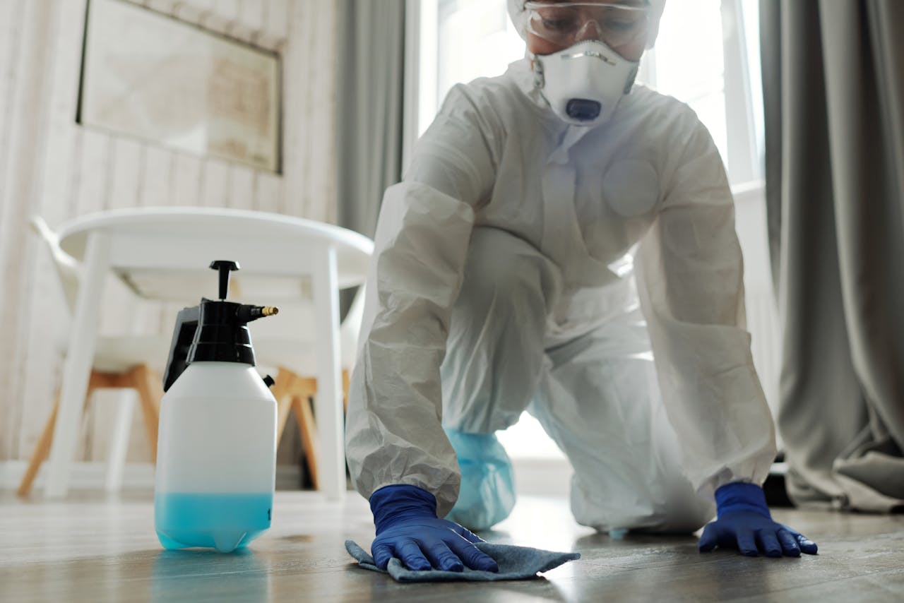 services-01 Person in protective gear cleaning a floor with disinfectant spray and cloth indoors.